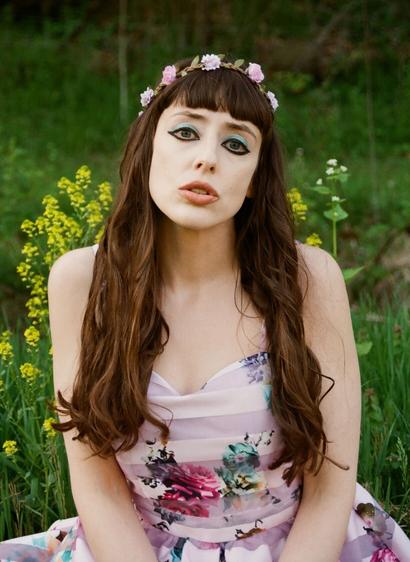 Author Gwendolyn Kiste, a fair-skinned woman with long, curled brown hair and an enigmatic expression. She wears a romantic pink flowered dress, a flower crown, and immaculate makeup. She sits in a field of flowers.