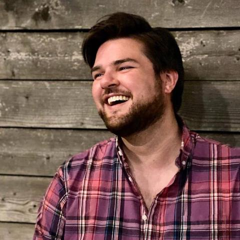 Author Sam Rebelein, a fair-skinned man with dark eyes, dark hair, and a beard. He wears a plaid shirt and is laughing happily. He stands in front of an unfinished wood wall.