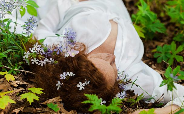 A close-up shallow depth of field image of a redheaded woman in a white flowy dress laying on the forest floor. Small lavender flowers of dames rocket and pale pink cranesbill ornament her hair.