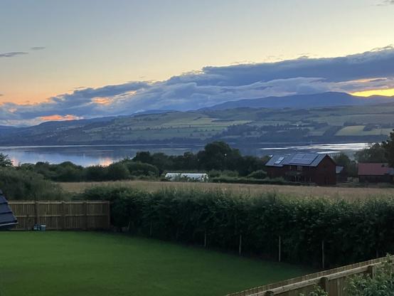 Very green lawn (not mine) in foreground with hedge at foot and wooden fence on left, then field of yellowing grass, trees, house and a polycrub on left. Glorious salmon, blue, gret white cloudy sky and Ben Wyvis range reflected in the very stll water of the Cromarty Firth.