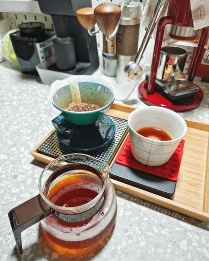 A coffee bar with grinders, manual espresso machine in the background. A cup, dripper and server with freshly brewed coffee.