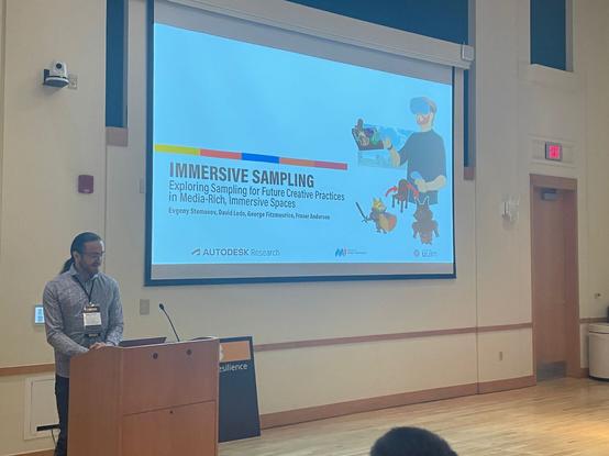 A person is standing behind a presenter's podium, looking downwards at a laptop computer. Behind and to the right of the person, a large projection screen is visible, showing a presentation slide. The slide reads "Immersive Sampling: Exploring Sampling for Future Creative Practices in Media-Rich, Immersive Spaces" and lists the following authors: Evgeny Stemasov,  David Ledo, George Fitzmaurice, Fraser Anderson, and a set of colored boxes, a graphical depiction, and several logos.