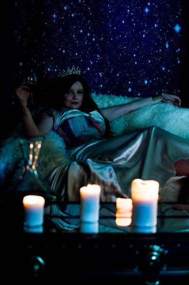 Darkly lit image of a white woman in a colorful corset and a silver dress, with dark hair and a pearly crown, lounging on a throne of furs against a backdrop of stars. She holds a glass of wine in her hand and lit candles sit on the mirrored table in front of her.
