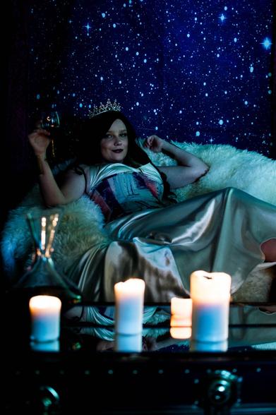 Darkly lit image of a white woman in a colorful corset and a silver dress, with dark hair and a pearly crown, lounging on a throne of furs against a backdrop of stars. She holds a glass of wine in her hand and lit candles sit on the mirrored table in front of her.