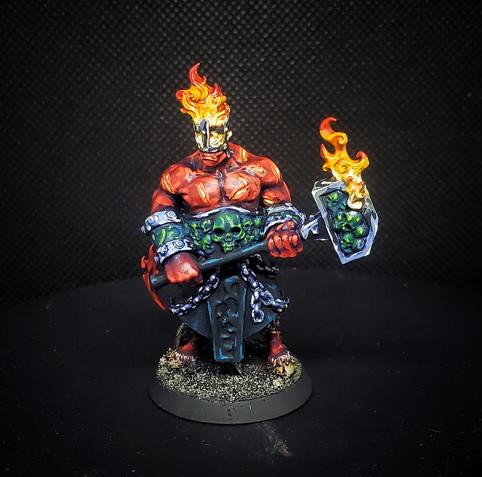 Photo of a painted miniature. An exceedingly buff red skinned devil with goat legs wearing a blue/black skirt and jade skull jewelry. He carries a large flaming hammer, and his head above the mouth is a brazier of burning fire.