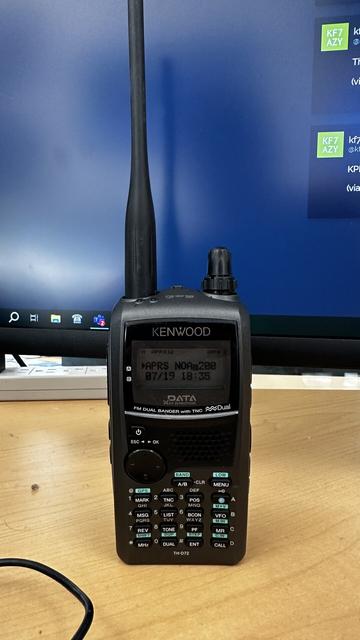 A handheld amateur radio with a number keypad on a wooden desk in front of a computer monitor.