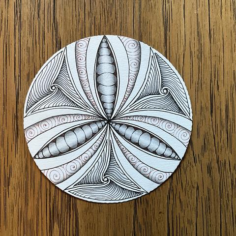 A piece of Zentangle art. On a white Zendala tile, this uses black and brown ink with graphite and white chalk shading. There’s a mix of orbs and spirals and waved lines creating a pea in the pod effect with bands and designs.