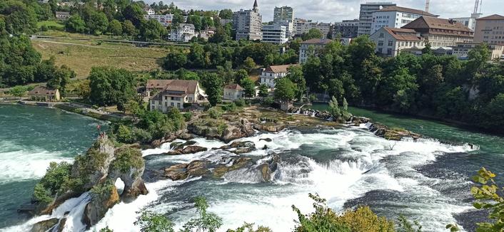 Kleine Wasserfälle, welches sich von einem Felsenriff in den Rhein stürzen. Im Hintergrund sieht man eine Stadt. Der Himmel ist klar und das Wetter gut.