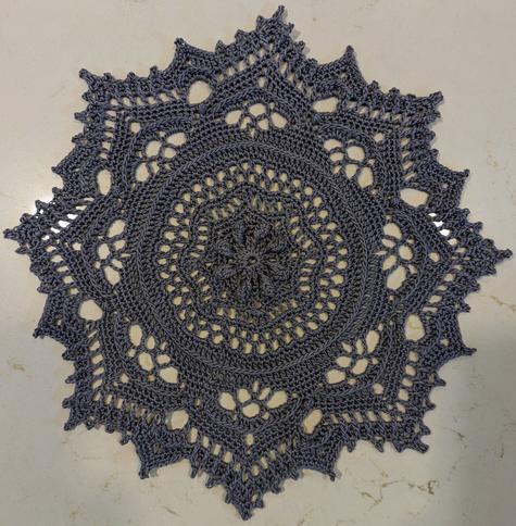 Silver colored thread lace crochet doily. The pattern is star/floral coming to 8 major points with minor points in between each point. It is textured and has raised circular and arched lines.