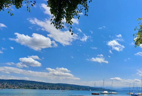 See, Segelboote, viel blauer Himmel mit weißen Wölkchen und in der Ferne Berge.