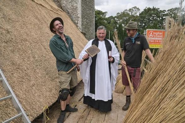Joyful scene of the robed vicar & two thatchers, all laughing, on a platform between the old thatch & the new reeds. The vicar is holding the legget or thatching bat.