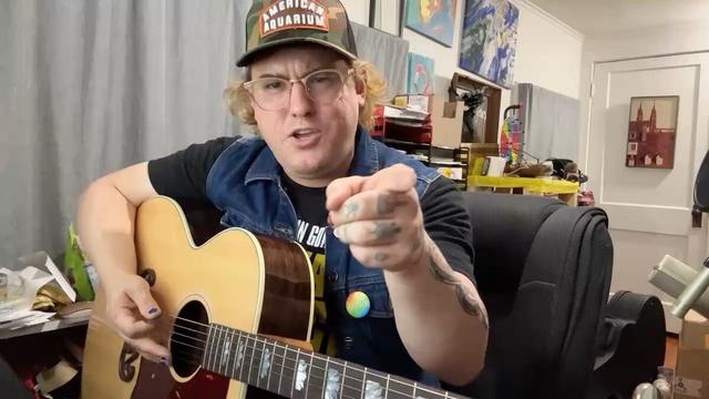 Adeem the Artist wearing a black tee shirt, denim vest and camouflage American Aquarium ball cap, introduces their new satirical Jason Andean “cover song” while sitting in an office chair in their house holding a guitar. Adeem is pointing at the camera with their left hand and ready to strum with their right.