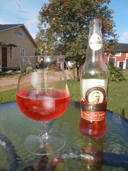 Lasipintaisella puutarhapöydällä iso lasi, jossa punaista siideriä ja jääpala sekä siideripullo, jossa osa siideristä vielä jäljellä. Taustalla keltainen vanha hirsirakennus, punainen vanha piharakennus, puu, nurtsia jne.