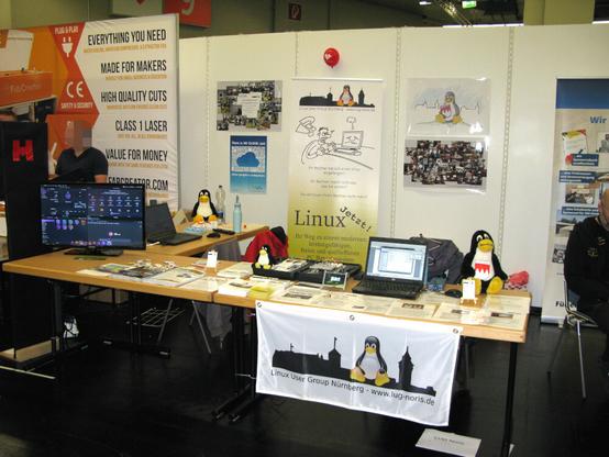 Auf dem Bild ist unser Messestand von 2022 zu sehen. Im Vordergrund zwei Tische mit unserem Prospektmaterial sowie einem Restbestand von Linux-Heften. Im Hintergrund an der Wand unser RollUp mit einigen Plakaten.