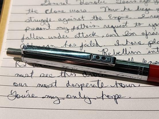 A retractable ball point pen sitting on a pad of paper with cursive writing. The pen top half is a silver metal. The metal pen clip is embossed with two hearts. The other half of the body of the pen is red. The text is the recorded "letter" that was recorded by Leia Organa into R2-D2 and addressed to Obi-Wan Kenobi during the beginning of the movie, Star Wars: A New Hope.