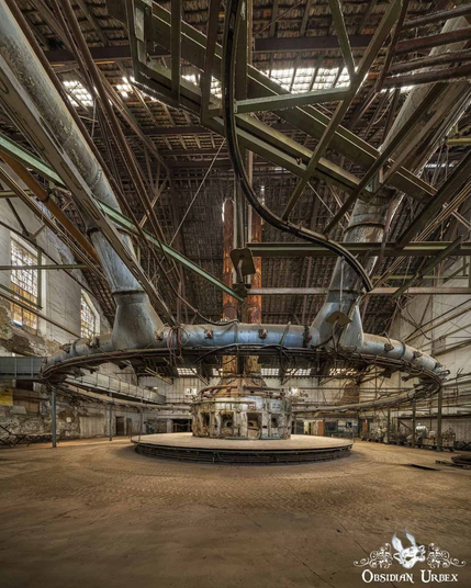 A huge rusty machine the middle of a room. There is a furnace in the middle, and a ring of pipes encircle it, hanging from the ceiling above
