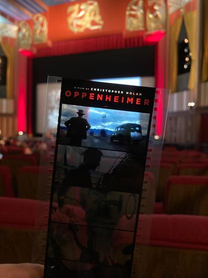 Photo in the theater getting ready to watch a 70mm print of Oppenheimer. i’m holding a novelty film strip given out to attendees of this screening.