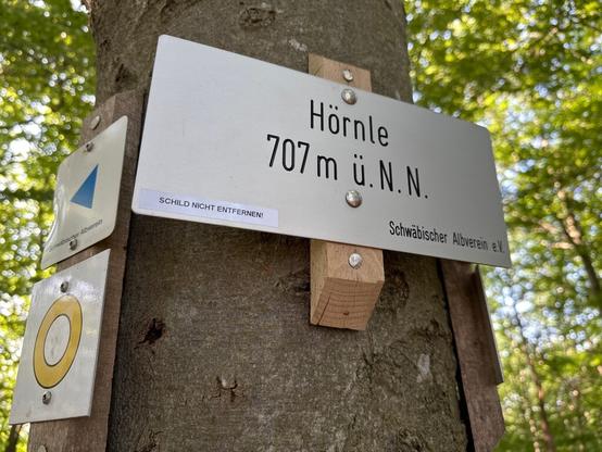 Schild auf einem Hügel mit dessen Namen „Hörnle“ und 700 Metern Höhe. Unten links ist ein Aufkleber angebracht auf dem „Schild nicht entfernen!“ steht.