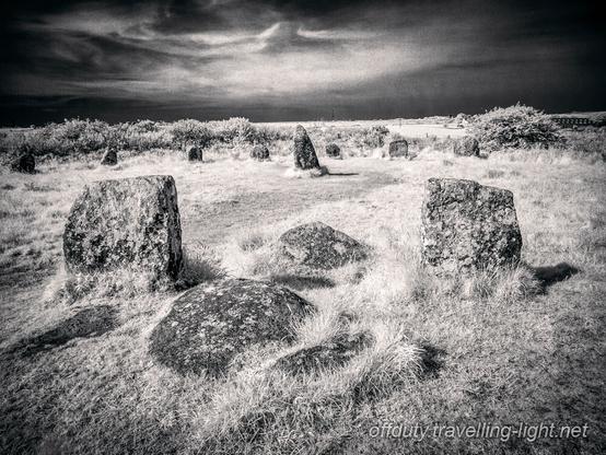 Black and white infrared photo of a stone circle in a grassy area, close up to a pair of stones, and a dark sky with wispy clouds.