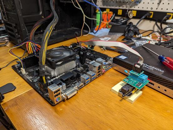 A photograph of a motherboard sitting on a workbench. It is connected to an off-camera power supply, and the CPU fan is running. Next to it is a mini-PC with a stack of little electronic bits sticking out of a USB port. A ribbon cable runs from this to a spring clip attached to a chip on the motherboard.