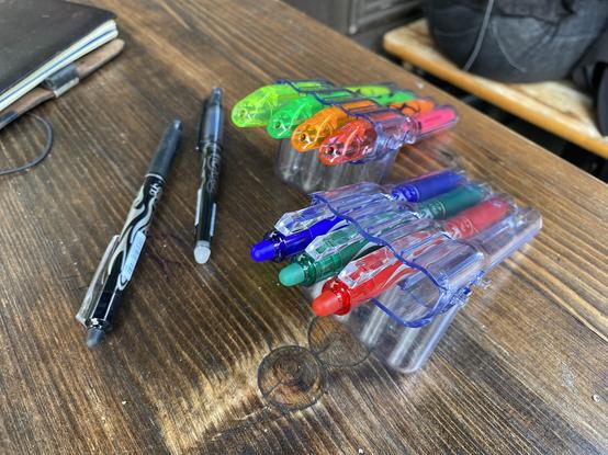 Two cases of Frixion pens (highlighters and coloured rollerballs) with the lid propping them up on a wooden table