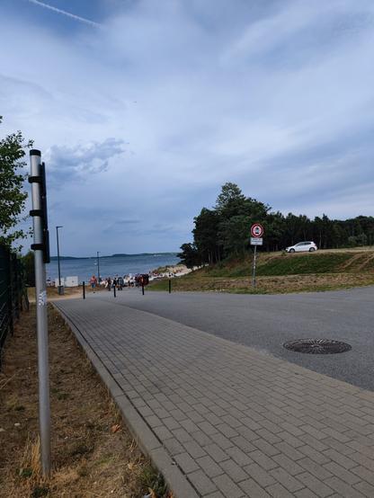 stromo opadająca asfaltowa droga w dół w kierunku plaży przy jeziorze, na pierwszym planie chodnik obok drogi