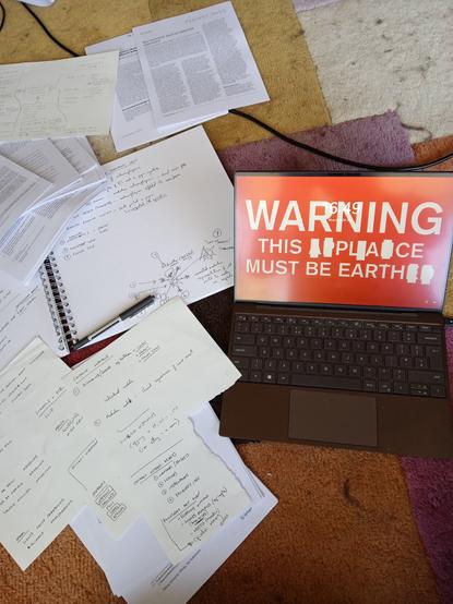 A large plain open notebook, with handwritten notes. Scraps of paper, also annotated. Research articles scattered on and under the papers. A pen, chewed at both ends. A laptop, open, showing a red screensaver with white text: WARNING, THIS PLACE MUST BE EARTH.

[The screensaver is adapted from an electrical appliance safety notice, and actually reads; Warning, this appliance must be earthed. Some letters have been overlaid with white boxes]
