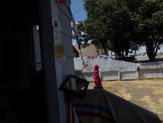 A string of panties (female) drapes out of the shaded West porch of a pacifist the panty peace flag goes over the yard and a low cut fence to a taller fence then runs to the right and is attached to a low branch of a tree over the fence.