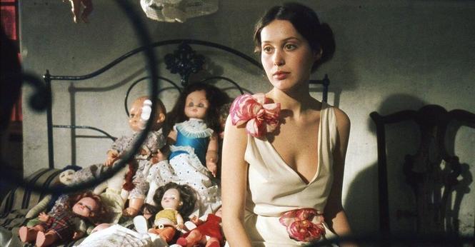 Prostitute María (Nadège Clair) sitting on her bed. The bed is covered in dolls.