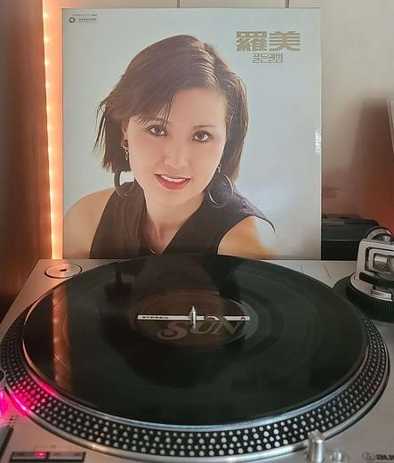 Image shows a turntable with a black vinyl record on the platter. Behind the turntable vinyl album outer sleeve is displayed. The front cover shows Na-Mi from the shoulders up looking at the camera.