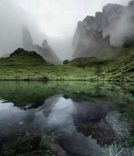 Two jagged, dog-toothed mountains almost completely enveloped in mist with lower, softer green-covered hills and a lake/pond reflecting them.