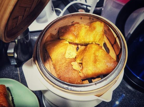 Three slices of apam balik (thick folded pancake with sweet filling) snugly placed in a bamboo dim sum steamer basket. The basket is on a multi-cooker that is boiling the water. To the top left of the photo, the steamer cover is moving out of frame (I was lifting it to take a photo of what's inside the basket).