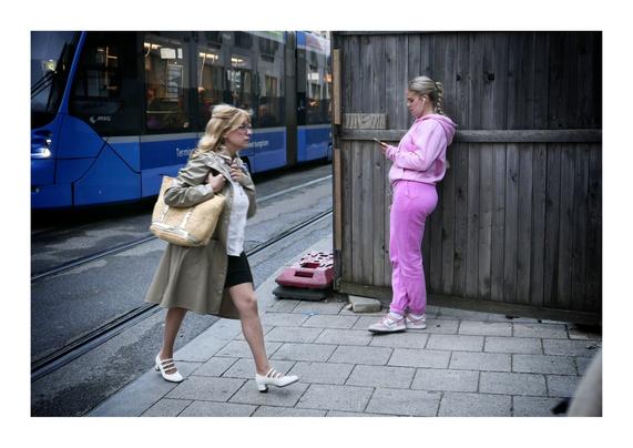 Zwei Frauen auf der Straße. Eine trägt einen knallpink Jogginganzug, lehnt sich gegen einen Bretterzaun und schaut in ihr Smartphone. Wahrscheinlich hat sie gerade den Film "Barbie" geschaut.
Die andere läuft mit einer großen Handtasche unter den Arm geklemmt vorbei. Sie trägt modische Kleidung.