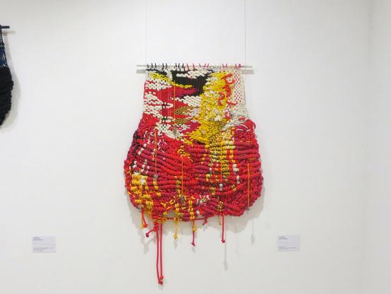 Alyona Repkina, “Hive”, 2023, textiles, steel whistles, 100 × 140 × 8 cm // An abstract knitted sculpture made of laces suspended from a horizontal stick. Dominant colours: red, yellow and crimson (or dark pink). Other colours: white and black. Eight steel whistles are suspended from the sculpture on yellow ropes, three above and five below (crypto “нет войне”/“net voine”/“no to war”?). There are no explanations on the label and near the sculpture whether or not you can touch the artwork. // Алёна Репкина, «Улей», 2023, шнурки, верёвки и стальные свистки, 100 × 140 × 8 см // Абстрактная вязаная скульптура из шнурков, подвешенная на горизонтальную палку. Доминирующие цвета: красный, жёлтый и малиновый (или тёмно-розовый). Другие цвета: белый и чёрный. К скульптуре на жёлтых верёвках подвешены восемь стальных свистков, три сверху и пять снизу («нет войне»?). На этикетке и возле скульптуры нет никаких пояснений, можно или нельзя прикасаться к работе.