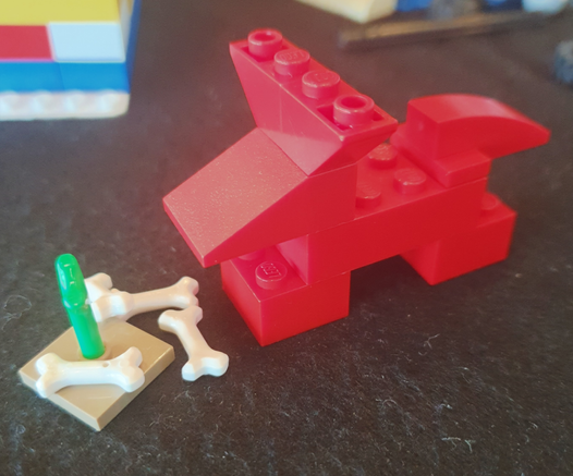 En figur bygget af røde LEGOklodser der svagt minder om en ræv, ved nogle kødben i LEGO og en grøn vækst.