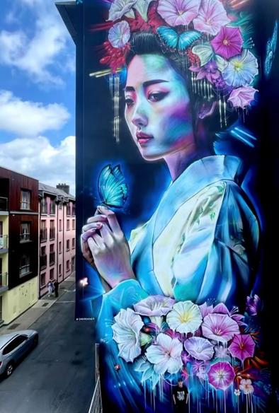 Streetartwall. On the outer wall of a multi-storey hotel is painted a huge mural of a beautiful geisha. On a black background she is depicted slightly tilted to the side. In bright colors, a lady in a white silk kimono can be seen holding a pink butterfly on her two hands. In her hair and on her forearm she wears a floral arrangement of orchids in pink, purple and white. Her gaze is not fixed on the butterfly. She is looking into the void.