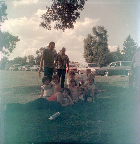 Slightly fogged color photo of a group in a park in front of cars which place the date at least 40 to 45 years ago.