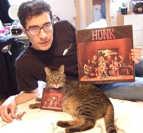 A picture of a man and his tabby cat reclining on a bed. The man holds a vinyl record-HONK— with little clay hippie figures of the band. The cat has a small CD version of the LP.