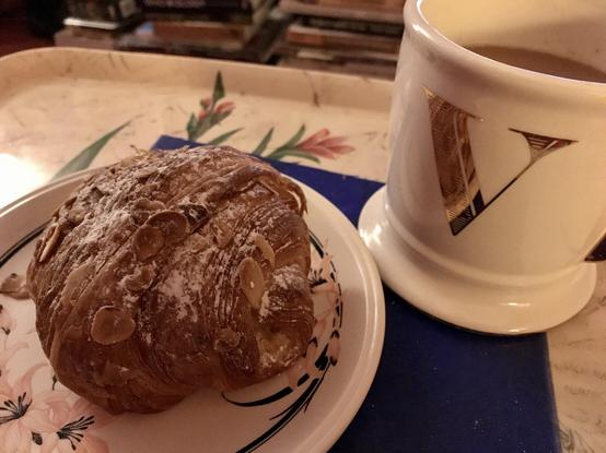 Photo of a large crumbly and flaky sort of round croissant, sitting on a plate beside a very large mug of coffee in a mug with a gold monogram V. Both mug and croissant are huge.