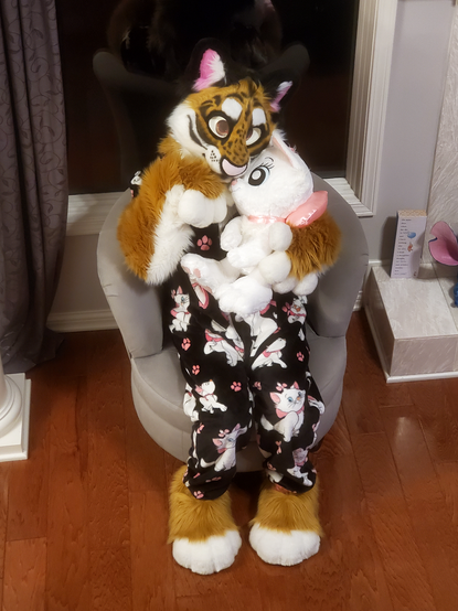 Fursuit photo. Demani is a smilodon which also known as a saber-toothed tiger. He is orange with white cheecks and fluff. He has small black stripes all around his face and two longer stripes under each eye. His ears are black with pink inside and white ear fluff. His nose is big and pink with black spots along his nose bridge. He is sitting in a chair, facing slightly to the right, left paw up at his chin, right paw hugging a large plush Marie from Disney's Aristocats. He is wearing black pajamas adored with various poses of the same Marie character. Marie is a white cat with blue eyes, pink inside ears, with a tiny pink bow on her head and a very large pink bow around her neck, with the bow part facing behind her.