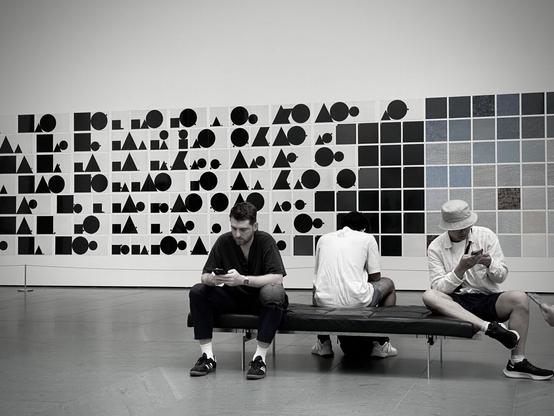 men sitting in a cushioned bench looking down sedentarily on their phones in front of art work at the museum of modern art in new york city