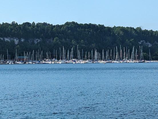 Sailboats parked in the distance on Georgian Bay with an escarpment behind them. It is sunny, and the water is a deep blue.