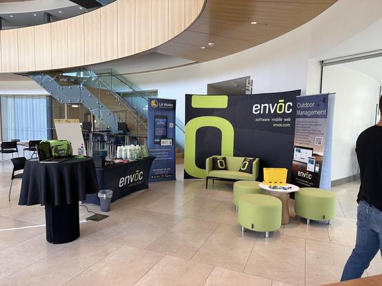 A large conference booth area laid out in a lounge setting with a couch, chairs, and espresso machine. In the background is a large backdrop with Envoc’s stylized “O” logo on it.