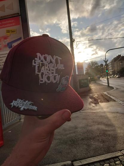 Bordeaux Rot farbene Snapback wird vor dem Sonnenuntergang bei bewölkten Himmel gehalten.
Auf der Cap steht "Don't Let The Label Label You", daneben ein gestrickter Hello Kitty Mermaid Pin.
Weil Sommer und so.

Im Schirm steckt ein Metall-Pin von der Band "Black Revelation".
