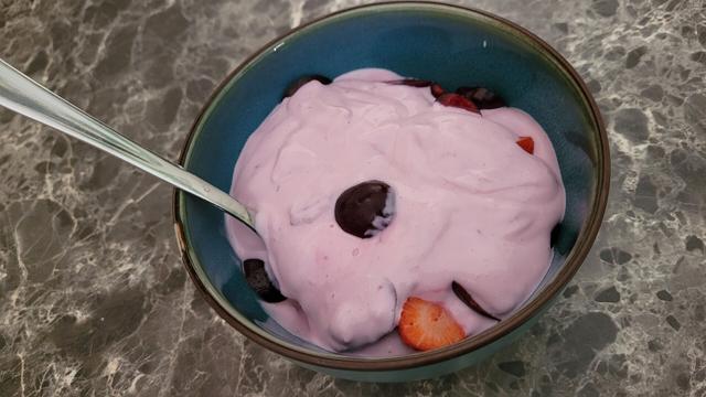 Een blauwe kom met roze-paars kleurige kwark erin en als toppings Aardbeien en Kersen
Ook zit er een metaalkleurige lepel in 
De achtergrond is een grijze marmerkleur