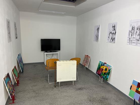 Im Hintergrund ein Fernseher, davor 2 Stühle (vor einer Heizung, die mitten im Raum steht). Am Boden rechts und links stehen Flanierbilder auf Flanierbildhaltern. An der Wand hängen verschiedene Use Cases für das Flanieren mit Flanierbildern.