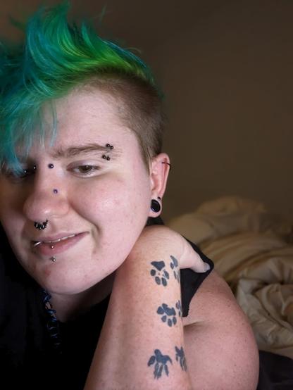 Selfie
Person with blue and green hair,  double eyebrow, a bridge, a high nostril, a septum, a vertical labret a lip ring, with tattooed paw prints on their arm.