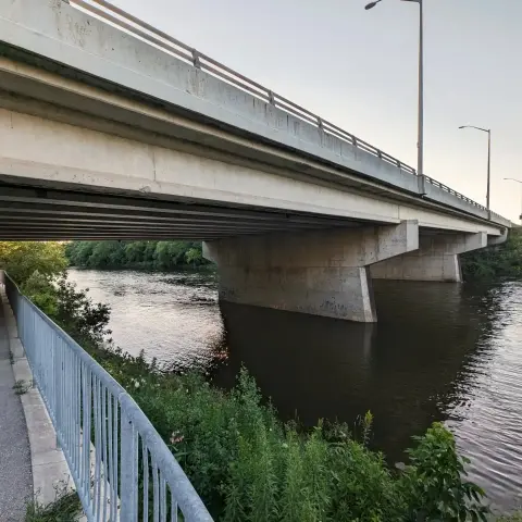 A picture taken from underneath a cement bridge that is crossing a river. There is a cycling path on the left, and some greenery lining the shore of the river.
