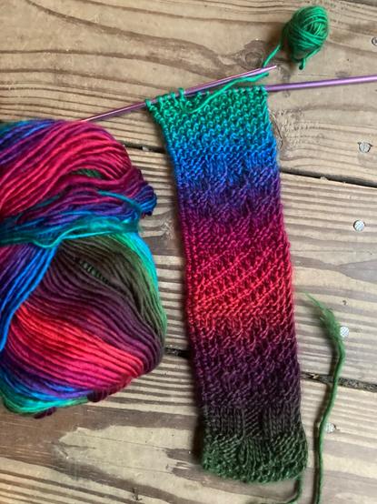 A knitting project and a skein of yarn sit on a wooden floor. I knit the reversible bookmark on under gauge needles (US 10 recommended, these are 6), with two stitches of seed for a side edge and four rows of garter (so two rows of pearls appear on each side) to separate my textured patterns. Seeing how slowly the colors change, I knew I had to make it wider than usual otherwise it’d go from green to dark green(!)