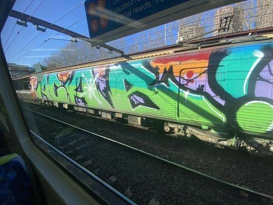 Graffiti mural covering an entire train carriage with the letters all capital saying ACAB! The photo is taken from inside a train carriage opposite the train with the graffiti. The lettering is different shades of green with a black outline. There are also some white highlights and sparkles painted on the letters. The train is in motion, pulling away from the platform, you can see the railway tracks, the overhead train wires, some skyscraper buildings and trees behind the train and you can see part of the train platform. The sky is partly visible too, it is a clear day and the sky is very blue. Colors used in the background of the graffiti are purple, orange and red.